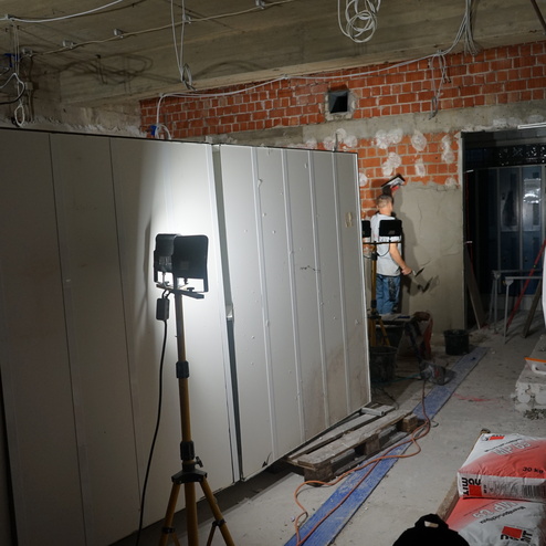 Handwerker bearbeitet eine Wand in einem Baustellenraum, beleuchtet von einem Stativstrahler; Baustellenmaterial liegt auf dem Boden.