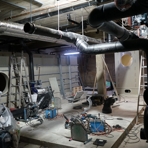 Bauinneres mit Rohren, Maschinen und Leitungen; eine Leiter steht an der Wand, Baustellenutensilien sind verstreut.