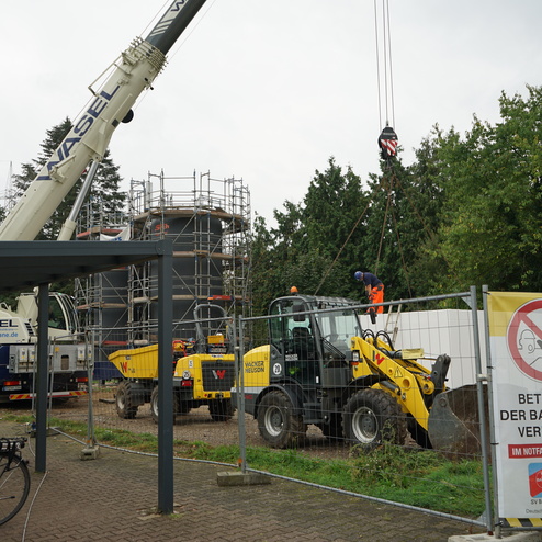 Baustelle mit Kr&auml;nen, Baggern und einem Arbeiter auf einer Leiter, umgeben von einem Bauzaun und Warnschildern.
