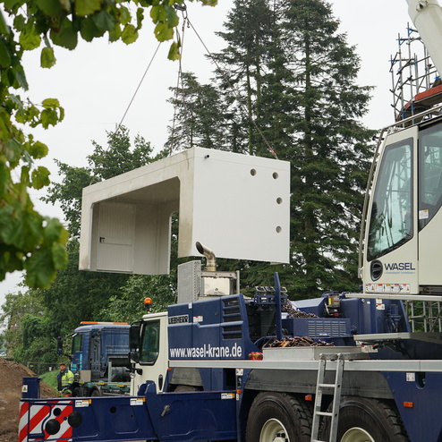 Kran hebt ein gro&szlig;es, rechteckiges Betonelement &uuml;ber einen Lkw auf einer Baustelle, umgeben von B&auml;umen.