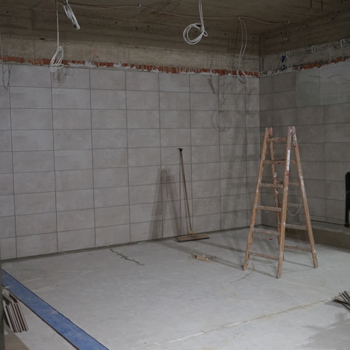 Blick auf einen unfertigen Raum mit gefliester Wand, einer Leiter und Baustellenutensilien auf dem Boden.