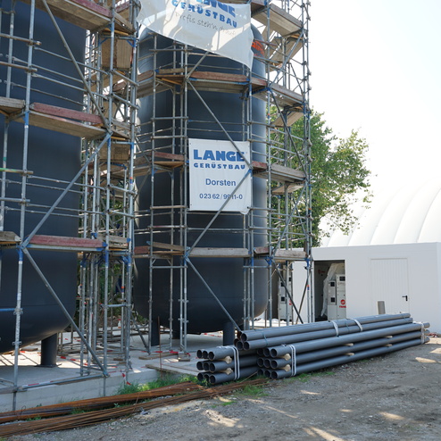 Bauger&uuml;st neben gro&szlig;en, runden Wassertanks, mit einem wei&szlig;en Baucontainer und gestapelten Rohren im Vordergrund.