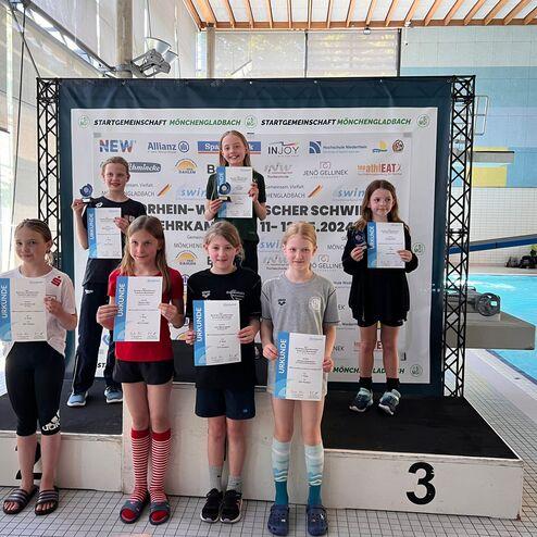 Siegerinnen einer Schwimmveranstaltung stehen auf dem Podest, halten Urkunden und Medaillen in einem Hallenbad.