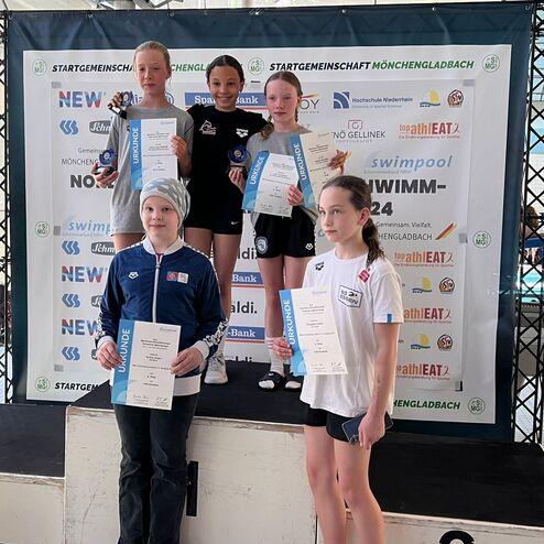 Fünf junge Schwimmerinnen posieren mit Urkunden und Medaillen auf einem Treppchen bei einem Wettkampf.