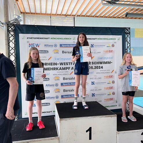 Drei Schwimmerinnen stehen auf dem Siegerpodest, halten Urkunden und Pokale beim Wettkampf in Mönchengladbach.