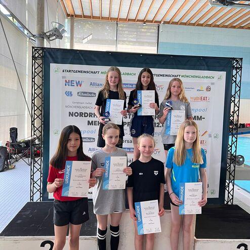 Gruppe von sieben Schwimmerinnen auf dem Siegertreppchen mit Urkunden und Medaillen in einem Hallenbad.