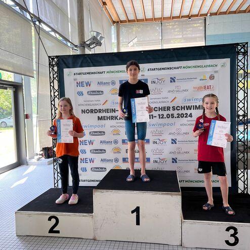 Drei Kinder stehen auf dem Podium nach einem Schwimmwettbewerb, halten Urkunden in der Hand und lächeln.