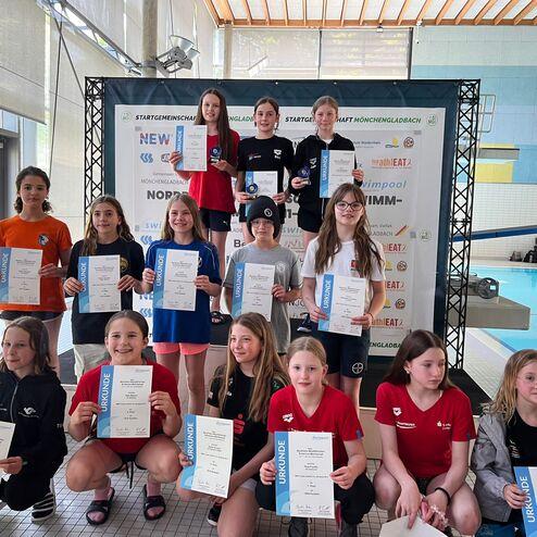Gruppenfoto von jungen Schwimmerinnen mit Urkunden auf dem Treppchen, im Hintergrund ein Schwimmbad.