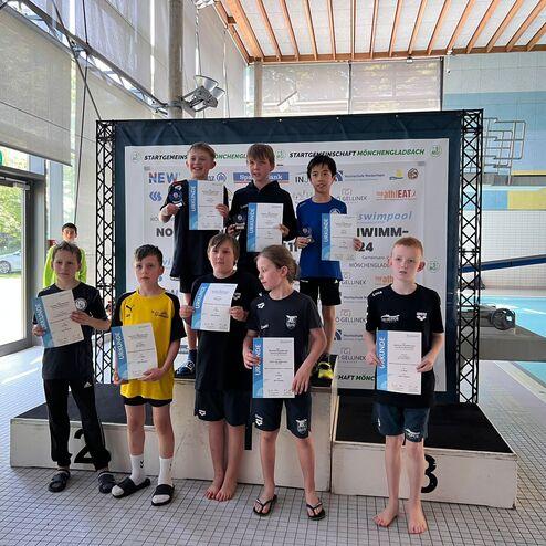 Kinder stehen auf einem Siegerpodest und halten Urkunden, im Hintergrund ein Schwimmbecken und Fenster mit Tageslicht.