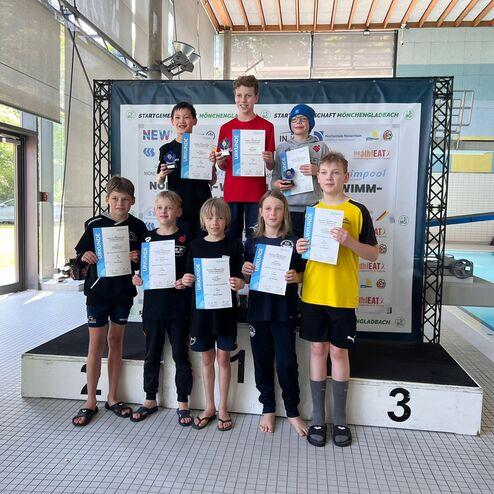 Gruppierung von sieben jungen Schwimmern auf dem Podium, die Urkunden und Medaillen in den Händen halten.