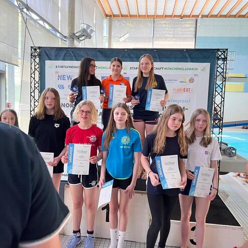 Gruppe von jungen Schwimmerinnen mit Urkunden auf einem Podium in einer Schwimmhalle.
