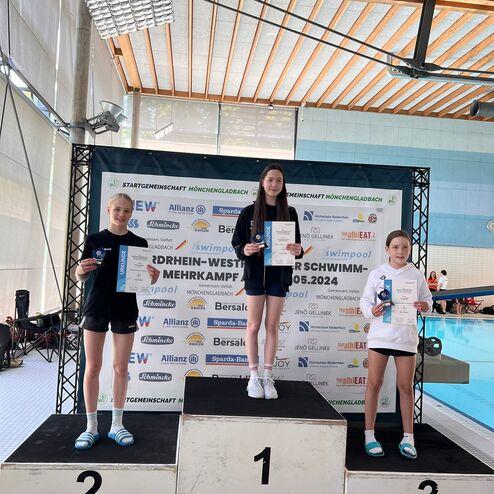 Drei Schwimmerinnen auf dem Siegertreppchen mit Urkunden und Medaillen bei einem Wettkampf in Mönchengladbach.