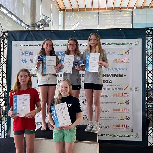 Vier Mädchen stehen auf einer Siegerehrung mit Urkunden und Medaillen in einem Schwimmbad. Hintergrund mit Wettkampfabzeichen.