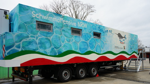 Bunter Trailer mit dem Schriftzug „Schwimmoffensive NRW“ und grafischen Wellenmotiven auf einem Parkplatz.