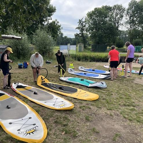 Gruppe von Personen, die auf einer Wiese mehrere Stand-Up-Paddle-Boards aufpumpen, im Hintergrund Bäume und ein Fahrzeug.