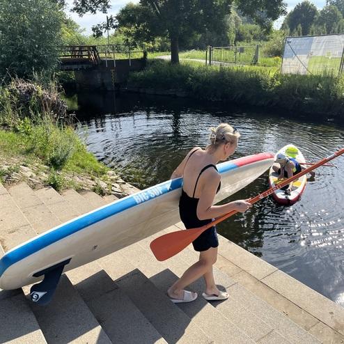 Frau trägt ein SUP-Board und paddel in Richtung Wasser, während ein Kind auf einem anderen Board sitzt.