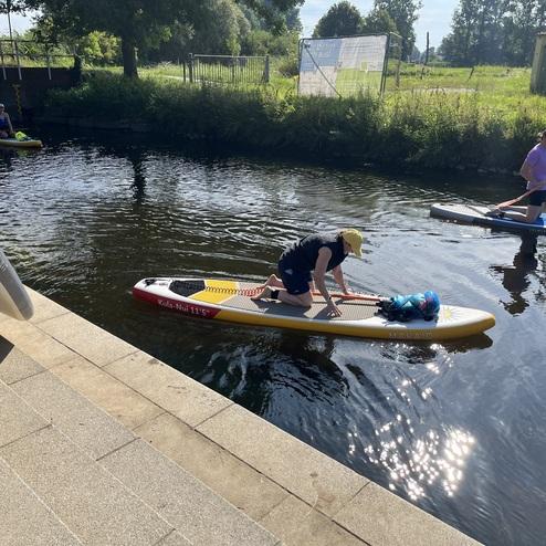Zwei Personen paddeln auf Stehpaddelbrettern auf einem ruhigen Gewässer an einer Betonstütze entlang.