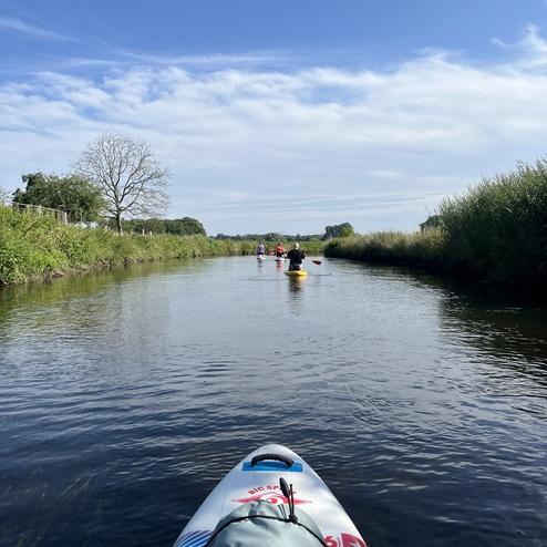 Blick von einem Stand-Up-Paddle-Board auf einen ruhigen Wasserweg mit Paddlern im Hintergrund und blauem Himmel.