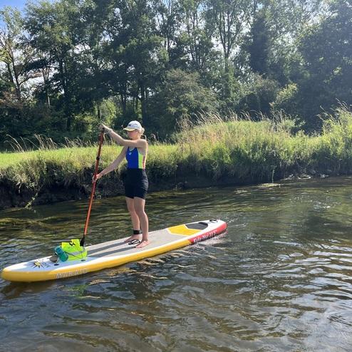 Frau paddelt auf einem Stand-Up-Paddle-Board im ruhigen Wasser, umgeben von grüner Ufervegetation und Bäumen.