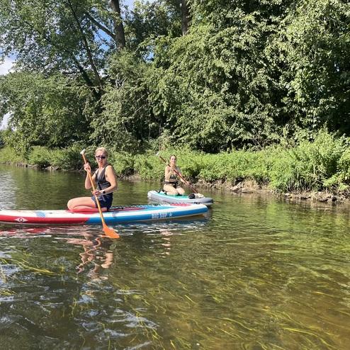 Zwei Frauen paddeln auf SUP-Boards in einem klaren Gewässer, umgeben von grüner Vegetation und einem Brückenbogen.