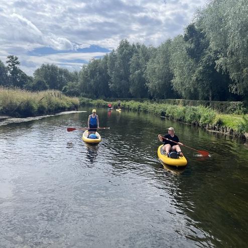 Zwei Personen paddeln auf gelben Kajaks auf einem ruhigen Gewässer, umgeben von üppigem Grün und bewölktem Himmel.