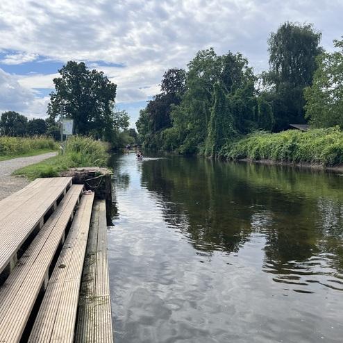 Ufer eines ruhigen Gewässers mit Holzstufen, umgeben von Bäumen und Sträuchern, im Hintergrund sind Ruderboote sichtbar.