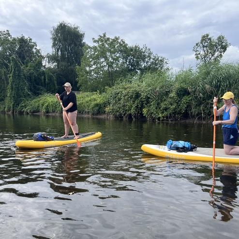 Zwei Frauen paddeln auf gelben Stand-Up-Paddleboards in einem ruhigen Gewässer, umgeben von grüner Ufervegetation.