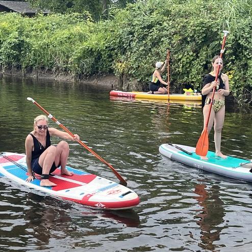 Zwei Frauen auf Stand-Up-Paddleboards in einem Fluss, umgeben von Pflanzen und einer weiteren Person im Hintergrund.
