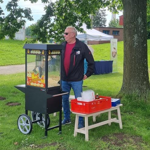 Mann steht neben einem Popcornwagen auf einer Wiese, daneben eine rote Kiste und ein Tisch.