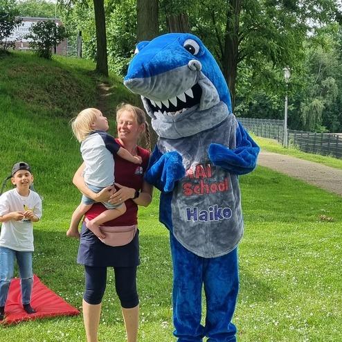 Mensch in Haifischkostüm steht mit Frau und Kleinkind auf einer Wiese, während ein Kind im Hintergrund spielt.