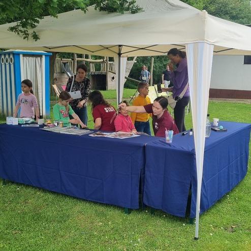 Sechs Kinder und zwei Betreuer stehen unter einem Zelt, während Gesichtsmalerei und Aktivitäten stattfinden.