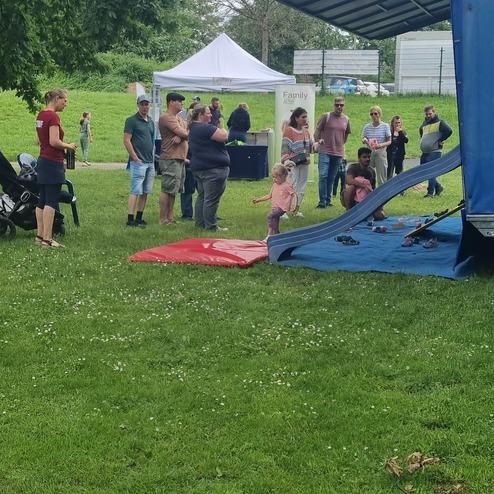 Gruppierung von Menschen, die vor einer blauen Rutsche im Freien stehen, umgeben von grüner Wiese und Bäumen.