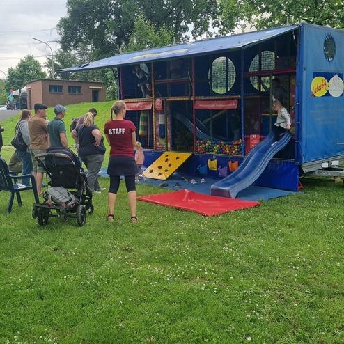 Gruppe von Menschen betrachtet einen bunten Spielanhänger mit Rutsche und Kletterelementen auf einer Wiese.