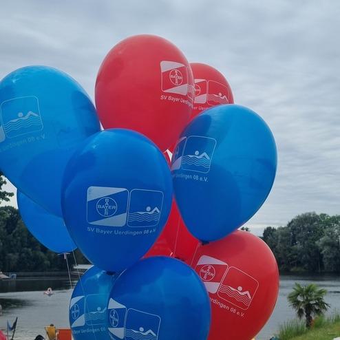 Bunte Luftballons in Rot und Blau mit dem Logo des SV Bayer Uerdingen 08 e.V. am Ufer eines Gewässers.