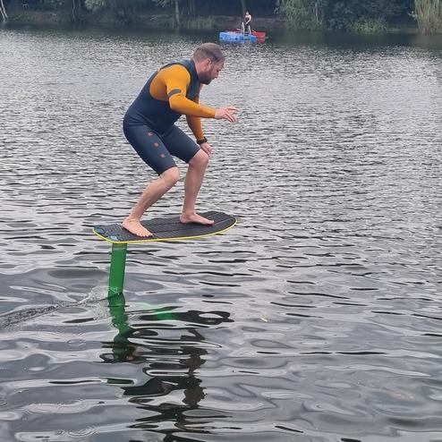 Mann auf einem Foilboard, der über dem Wasser steht, während im Hintergrund eine Person in einem Boot paddelt.