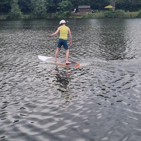 Person im gelben Schwimmwesten steht auf einem Paddleboard auf einem ruhigen Gewässer, umgeben von Bäumen.