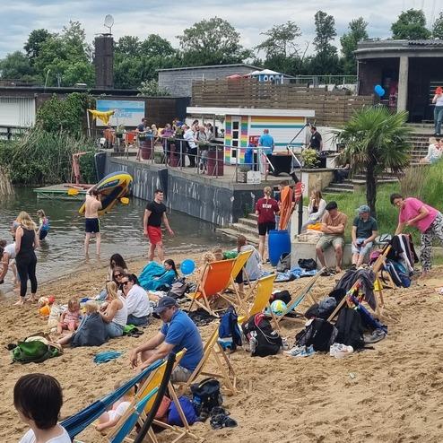 Bunte Liegestühle stehen am Sandstrand, während Menschen am Wasser und auf der Terrasse entspannen und spielen.