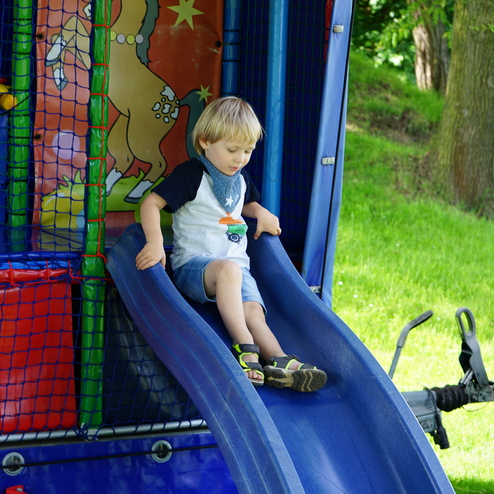 Ein Junge mit blonden Haaren rutscht auf einer blauen Rutsche in einem Spielbereich mit buntem Hintergrund.