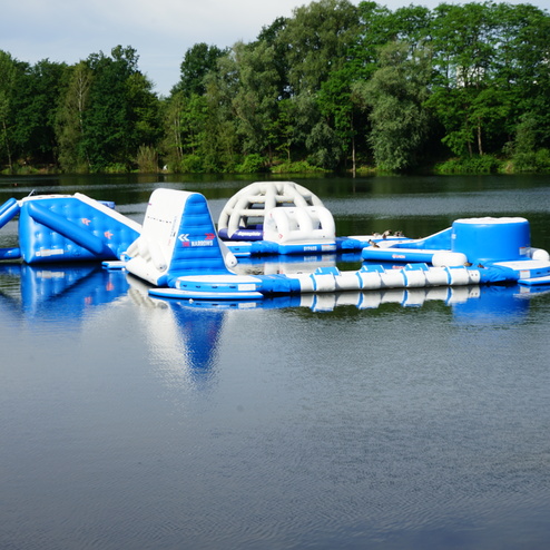 Schwimmende, luftgefüllte Spielgeräte in Blau und Weiß auf einem ruhigen See, umgeben von grünen Bäumen.