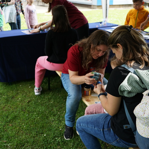 Zwei Frauen sitzen auf Bänken, während eine Frau einem Kind eine handwerkliche Aktivität zeigt. Um sie stehen mehrere Kinder.