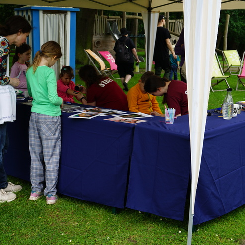 Zwei Kinder und eine Erwachsene betrachten Bilder an einem blauen Tisch unter einem Zelt auf einer Wiese.