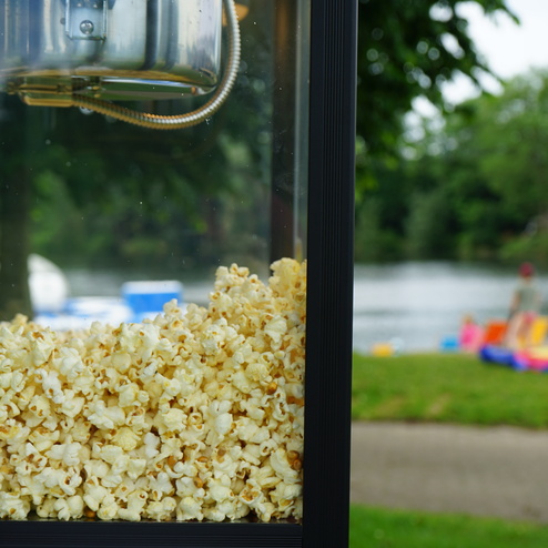Nahaufnahme einer Popcornmaschine mit frischem Popcorn, im Hintergrund ein Gewässer und Menschen beim Entspannen.