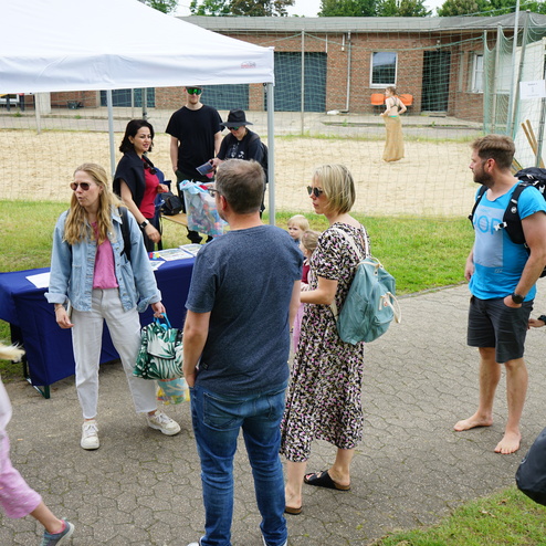 Menschengruppe bei einem Event unter einem Zelt, mit Informationsständen und einem Sandplatz im Hintergrund.
