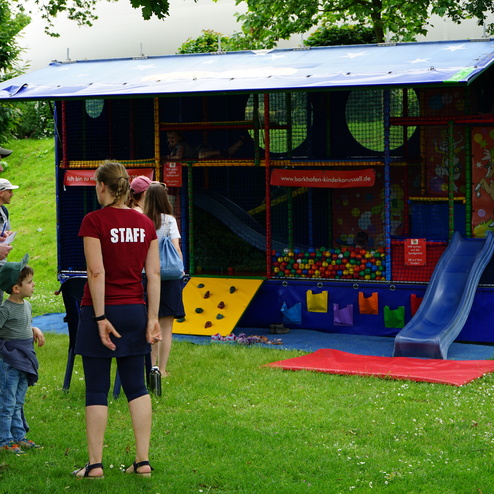 Familien stehen vor einem bunten Spielhaus mit Rutsche und Bällen, Mitarbeiterin in rotem Shirt erklärt das Angebot.