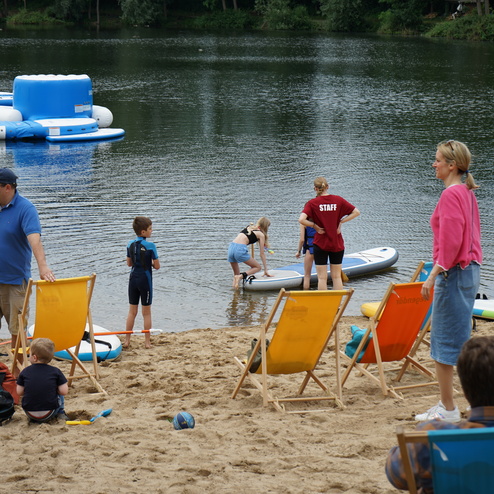 Menschen stehen am Ufer eines Sees, einige nutzen Stand-Up-Paddles, während andere am Strand entspannen.