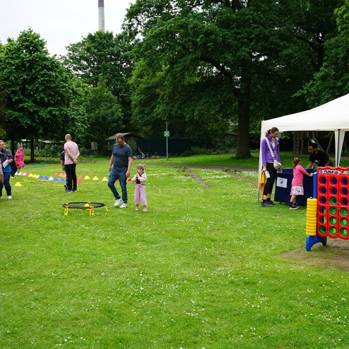 Familien spielen im Park, umgeben von Bäumen und grüner Wiese, mit einem Zelt und verschiedenen Spielstationen.