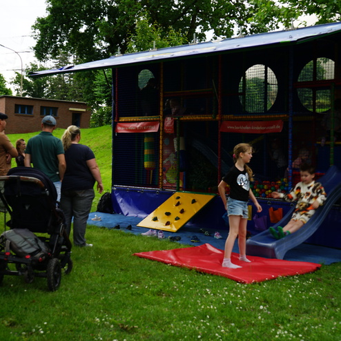 Kinder und Erwachsene stehen vor einem Spielmobil mit Rutsche und Bällen auf einer Wiese. Ein Kind rutscht gerade herunter.