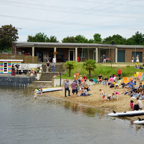 Menschen entspannen am Sandstrand eines Badesees, mit Sonnenliegen und Wassersportgeräten im Hintergrund.