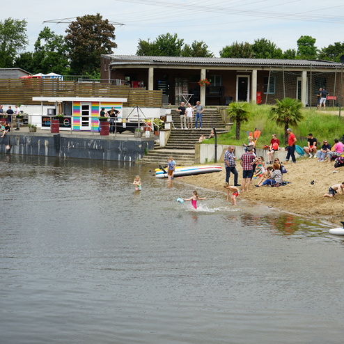 Blick auf einen Strand mit Menschen, die im Wasser spielen, neben einem Gebäude und mehreren Stand-Up-Paddleboards.