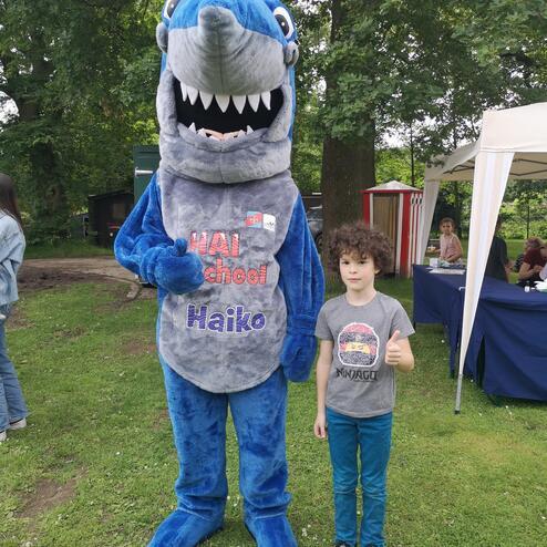 Kind mit Daumen hoch steht neben einem blauen Haifisch-Maskottchen auf einer Wiese, umgeben von Bäumen.
